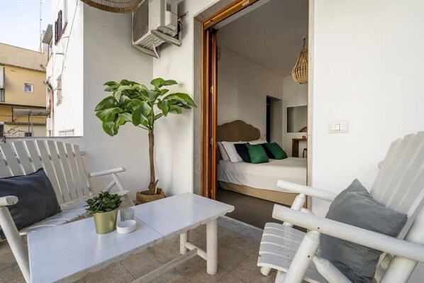 A balcony with a white table, two white chairs, a potted plant, and a bed with green pillows in the background. - Morada 181 B&B (Bari)