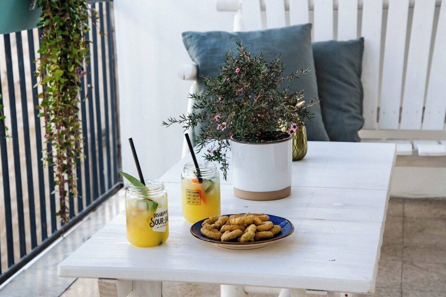 Una mesa blanca con dos bebidas, un plato de aperitivos y una planta en maceta.