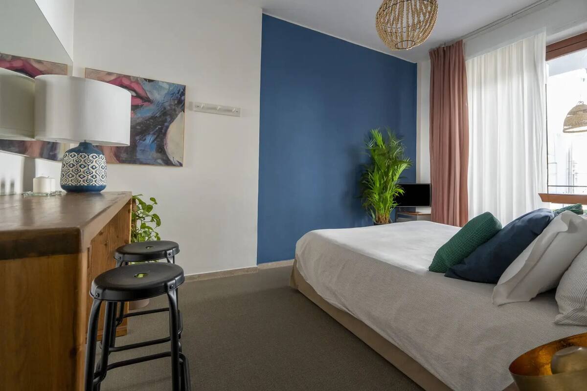 A bedroom with a bed, a desk, two stools, a potted plant, and a painting on the wall.
