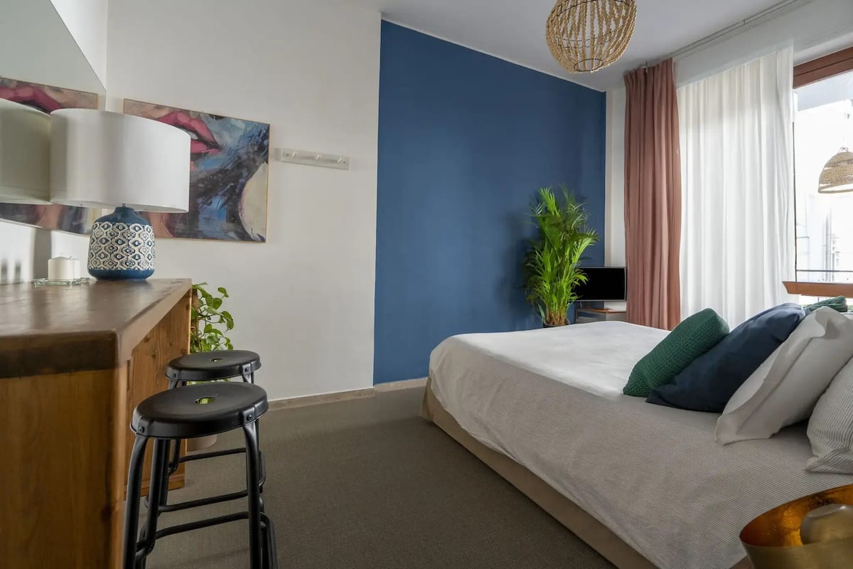 A bedroom with a bed, a desk, two stools, a potted plant, and a painting on the wall.