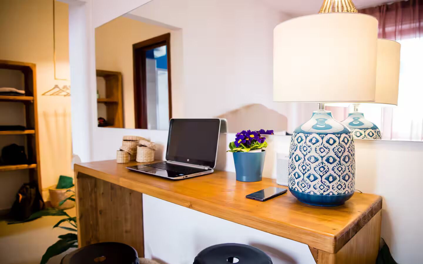 A wooden desk with a laptop, a smartphone, a potted plant, and a patterned lamp.