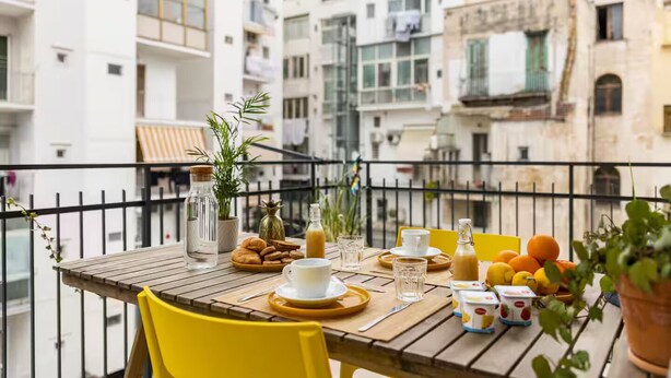 Une table en bois, entourée de chaises jaunes, avec une bouteille d’eau, un verre de jus de fruits, une assiette de pâtisseries et une tasse de café, le tout placé sur un balcon d’où l’on peut admirer des immeubles à plusieurs étages.
