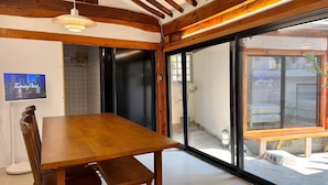 Un coin repas avec une table et des chaises en bois, un téléviseur fixé au mur et de grandes portes-fenêtres coulissantes donnant sur un espace extérieur.