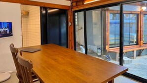 Une table à manger en bois, des chaises, un téléviseur fixé au mur et une porte-fenêtre coulissante donnant sur un espace extérieur avec un banc.