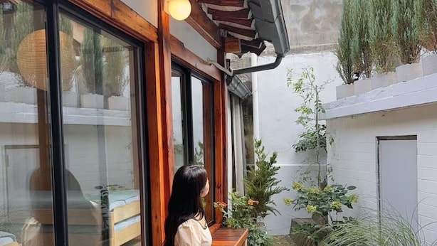 Une femme est assise sur un banc en bois dans une cour intérieure dotée de grandes fenêtres, de plantes en pot et offrant une vue sur un bâtiment en arrière-plan.