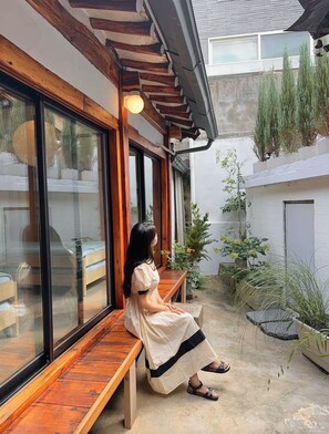 Une femme est assise sur un banc en bois dans une cour intérieure dotée de grandes fenêtres, de plantes en pot et offrant une vue sur un bâtiment en arrière-plan.