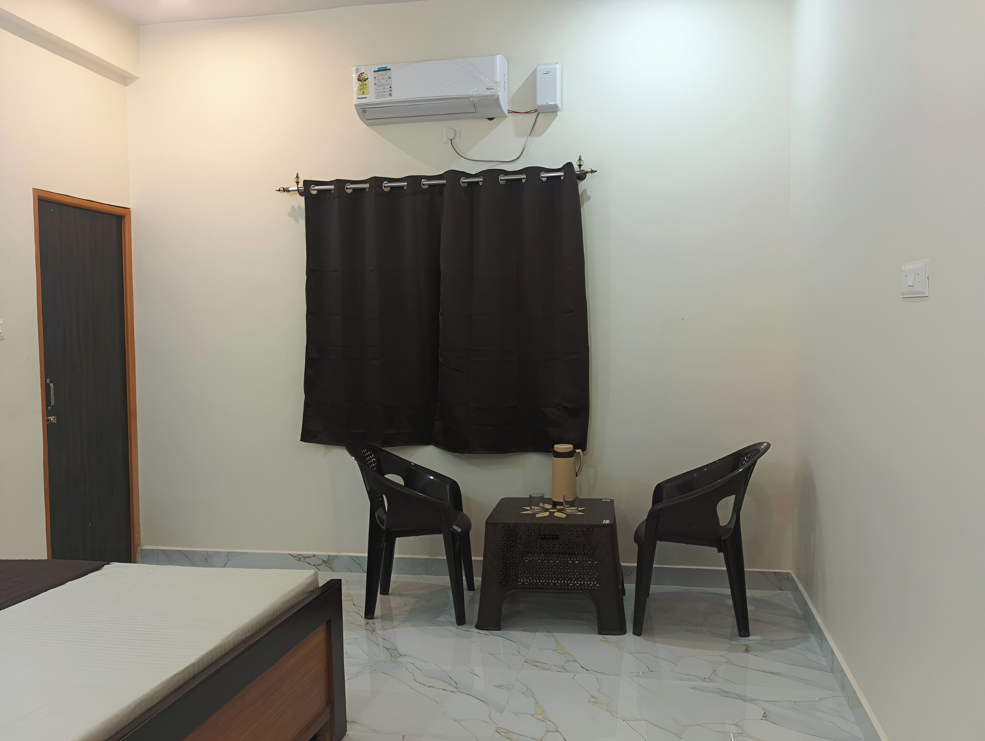 A small room with a bed, two chairs, a table, a curtain, and an air conditioning unit.
