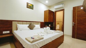 A neatly made bed with white linens and a headboard, a small bedside table with a lamp, a wooden wardrobe, and a bathroom visible through an open door. - Hotel Babylon Elegance (Mathura)