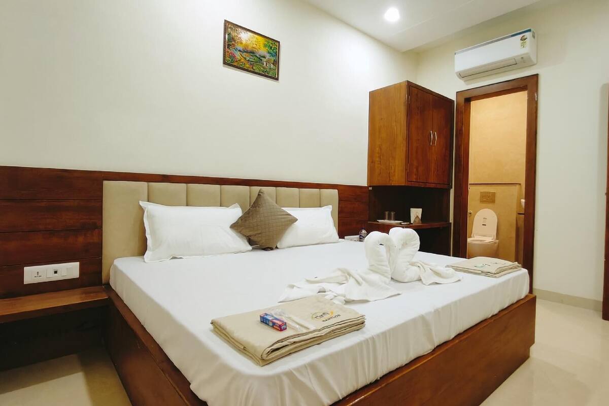 A neatly made bed with white linens and a headboard, a small bedside table with a lamp, a wooden wardrobe, and a bathroom visible through an open door.