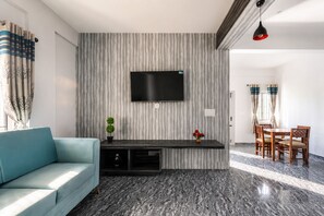 A modern living room with a flat-screen TV mounted on a textured wall, a light blue sofa, a small plant, and a dining area with wooden furniture. - Indigo Studio by Jadecaps 1bhk New Town Kolkata (Barasat)