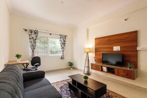 Uma sala de estar com sofá, mesa de centro, televisão sobre um armário de madeira, abajur e janela com cortinas.