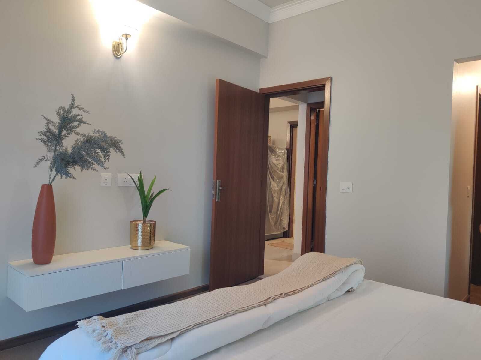 Une chambre avec un lit, une table de chevet, une étagère fixée au mur avec une plante, et une porte donnant sur une autre pièce.