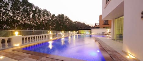 Une piscine dotée d’une balustrade en verre, entourée d’une terrasse dallée et bordée d’un bâtiment aux grandes fenêtres.