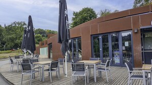 Een terras met tafels en stoelen, een houten terras en een modern gebouw met grote ramen.