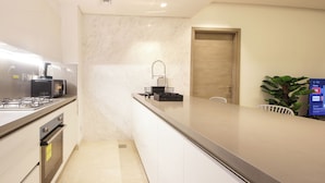 A modern kitchen with a built-in microwave, gas stove, and oven. There is a sink with a faucet and a countertop with a coffee machine. A wooden door and a plant are visible in the background. - 1 BR Burj Khalifa View Sterling East (Dubai)