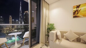 A modern living room with a sofa and a small table, large windows with a city view, and a painting on the wall. - 1 BR Burj Khalifa View Sterling East (Dubai)
