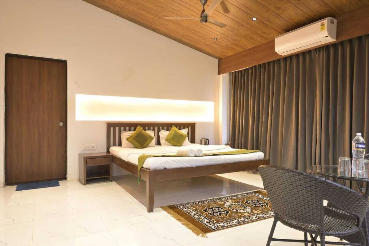 A modern hotel room with a wooden ceiling, a bed with green and white bedding, a bedside table, a glass table with chairs, a large window with curtains, and an air conditioning unit.