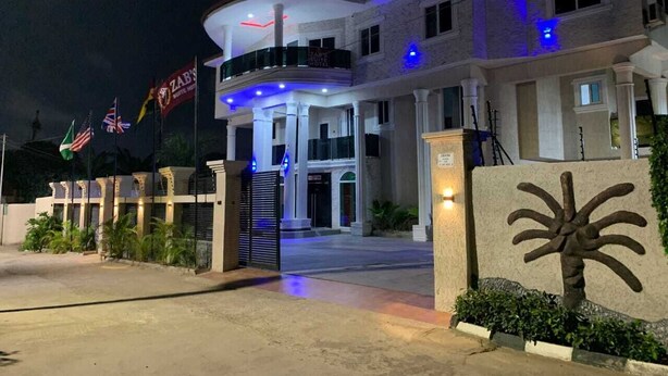 A hotel entrance with a large, curved facade, multiple flags, and a prominent palm tree sculpture. - Zab's Suite Hotel  (Accra)