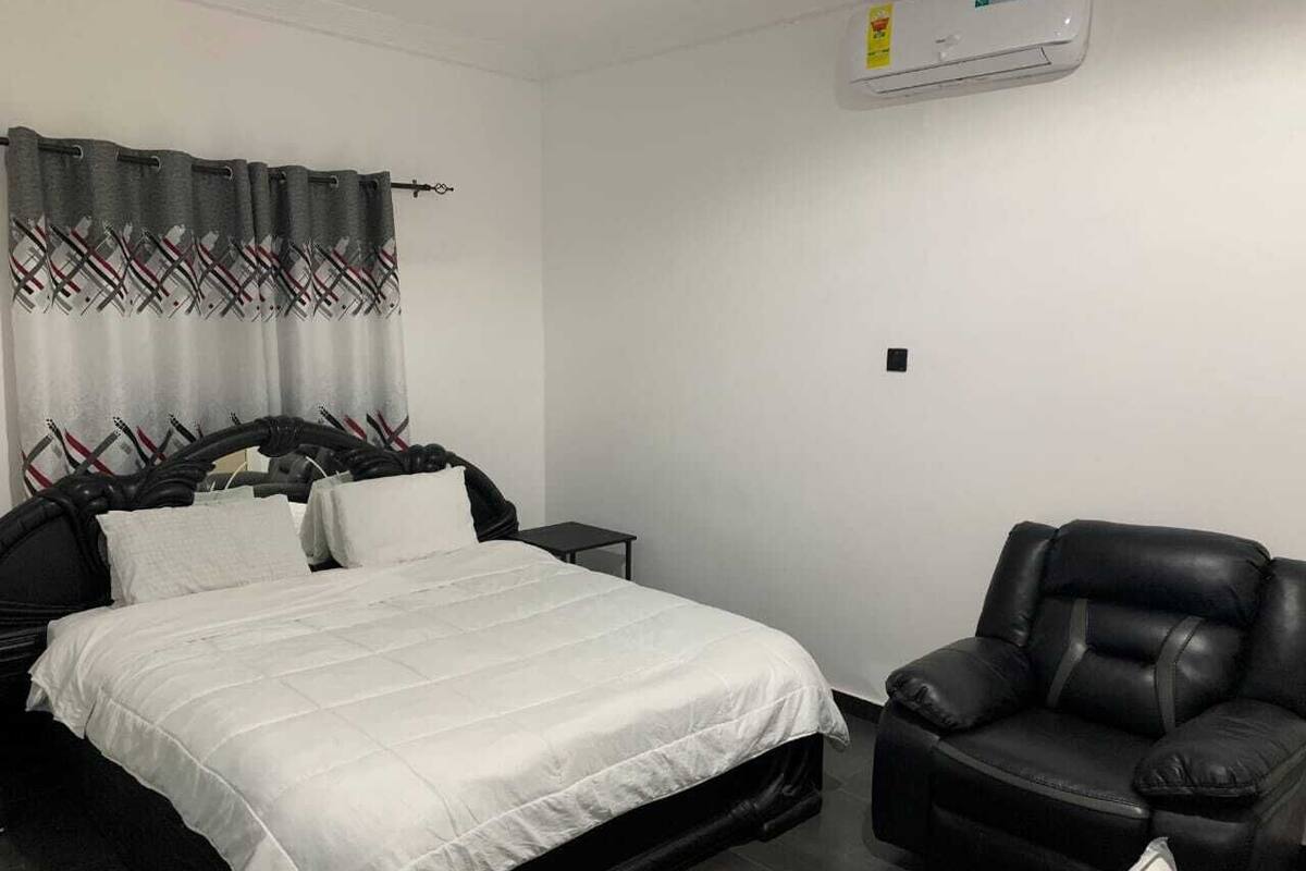 A bedroom with a bed, a chair, a nightstand, and a wall-mounted air conditioner.