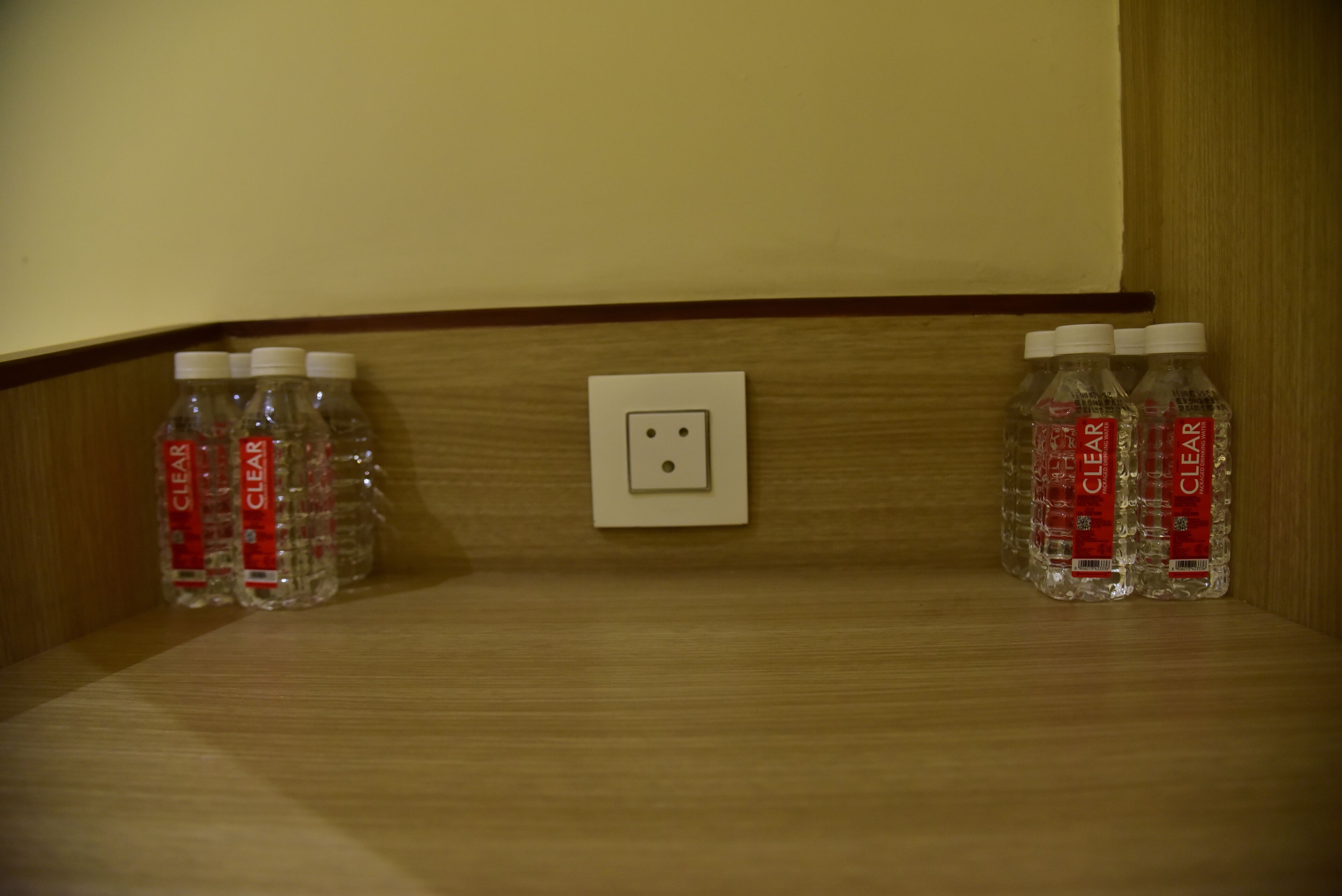 A wooden shelf with a white electrical outlet and two rows of clear plastic water bottles with red labels.