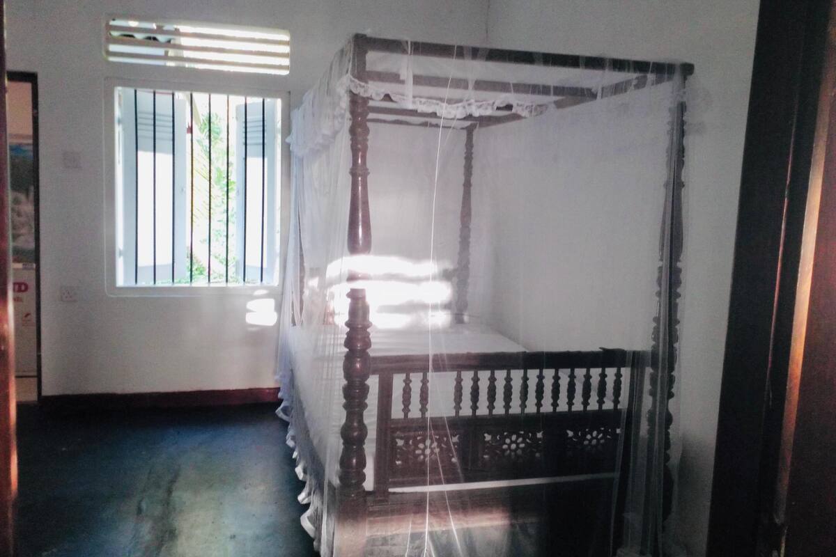A room with a four-poster bed, a window with vertical blinds, and a blue carpet.