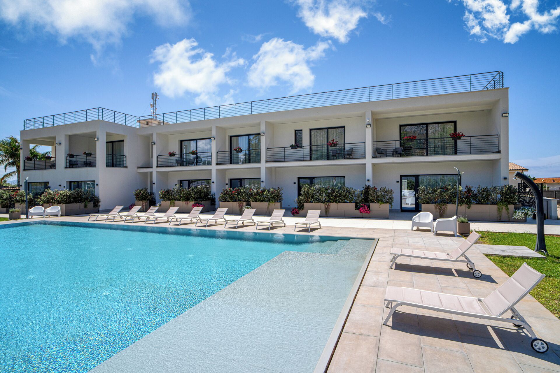 A modern multi-story hotel with a swimming pool, lounge chairs, and balconies with plants.