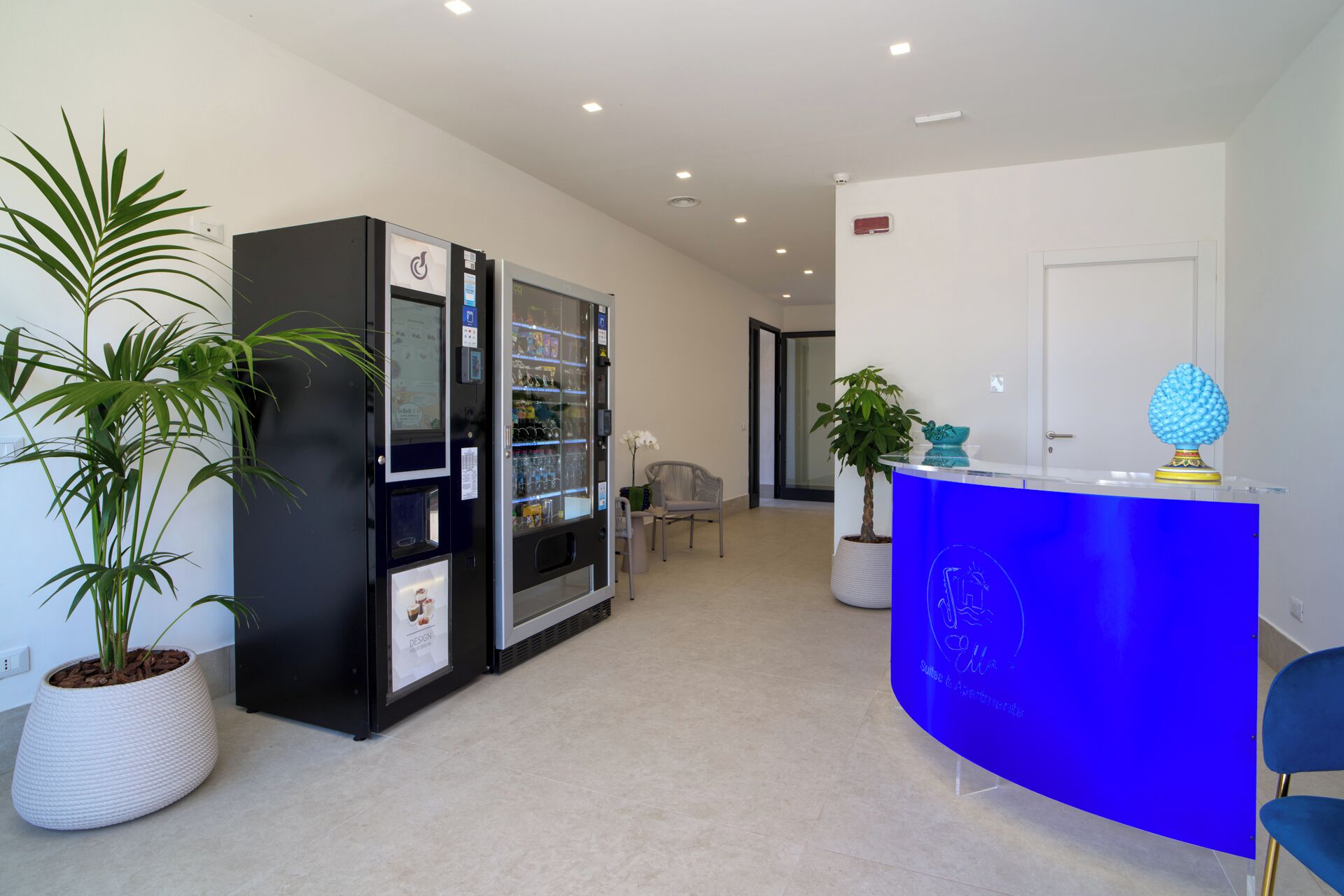 A reception area with a blue counter, a potted plant, a seating area with a chair, and two vending machines.