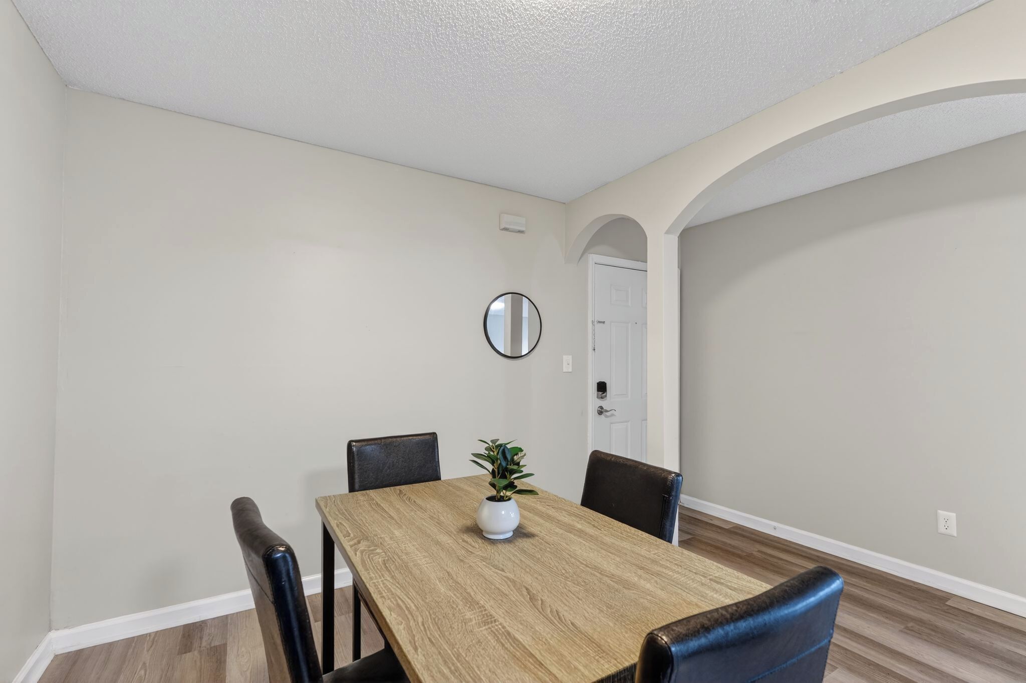 Un comedor con una mesa de madera, cuatro sillas, una planta en maceta y un espejo redondo en la pared.