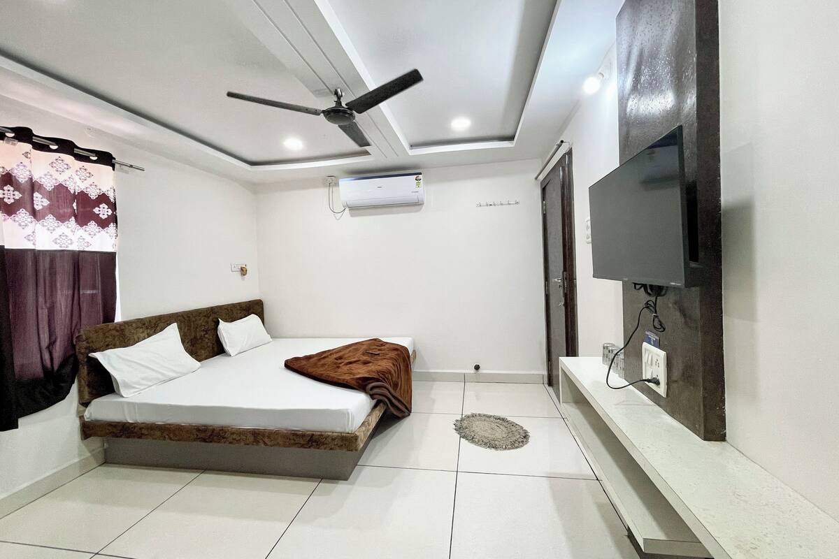 A bedroom with a bed, a ceiling fan, an air conditioning unit, a TV mounted on the wall, and a bedside table.