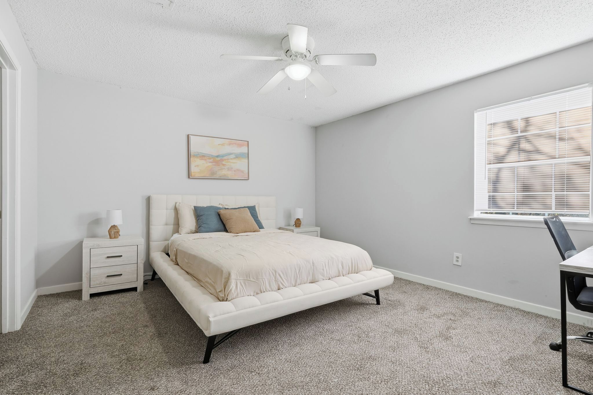 A bedroom with a bed, nightstands, a ceiling fan, a window with blinds, and a painting on the wall.