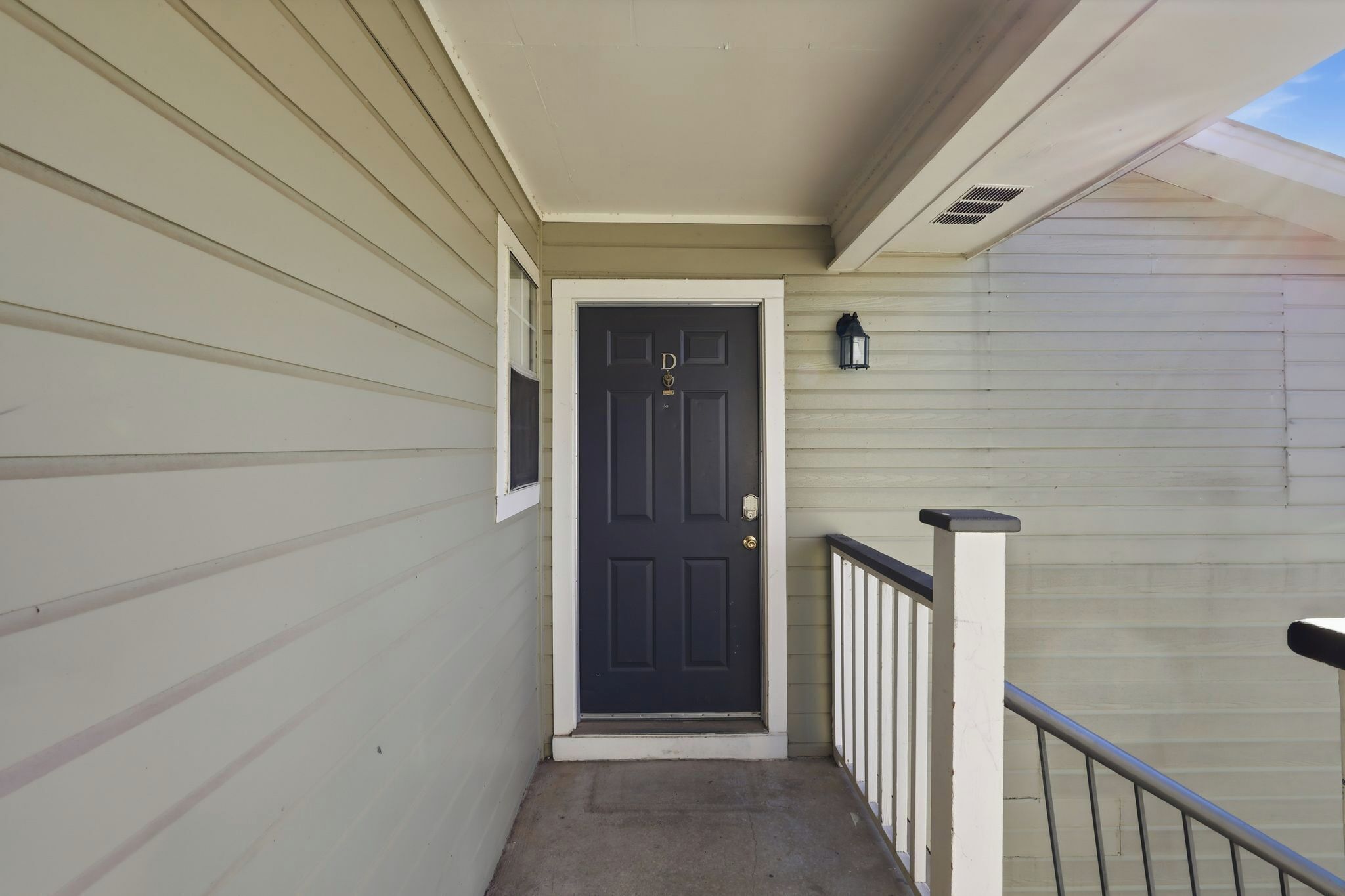 A narrow, covered exterior corridor with a dark door, a small window, and a wall-mounted light fixture.