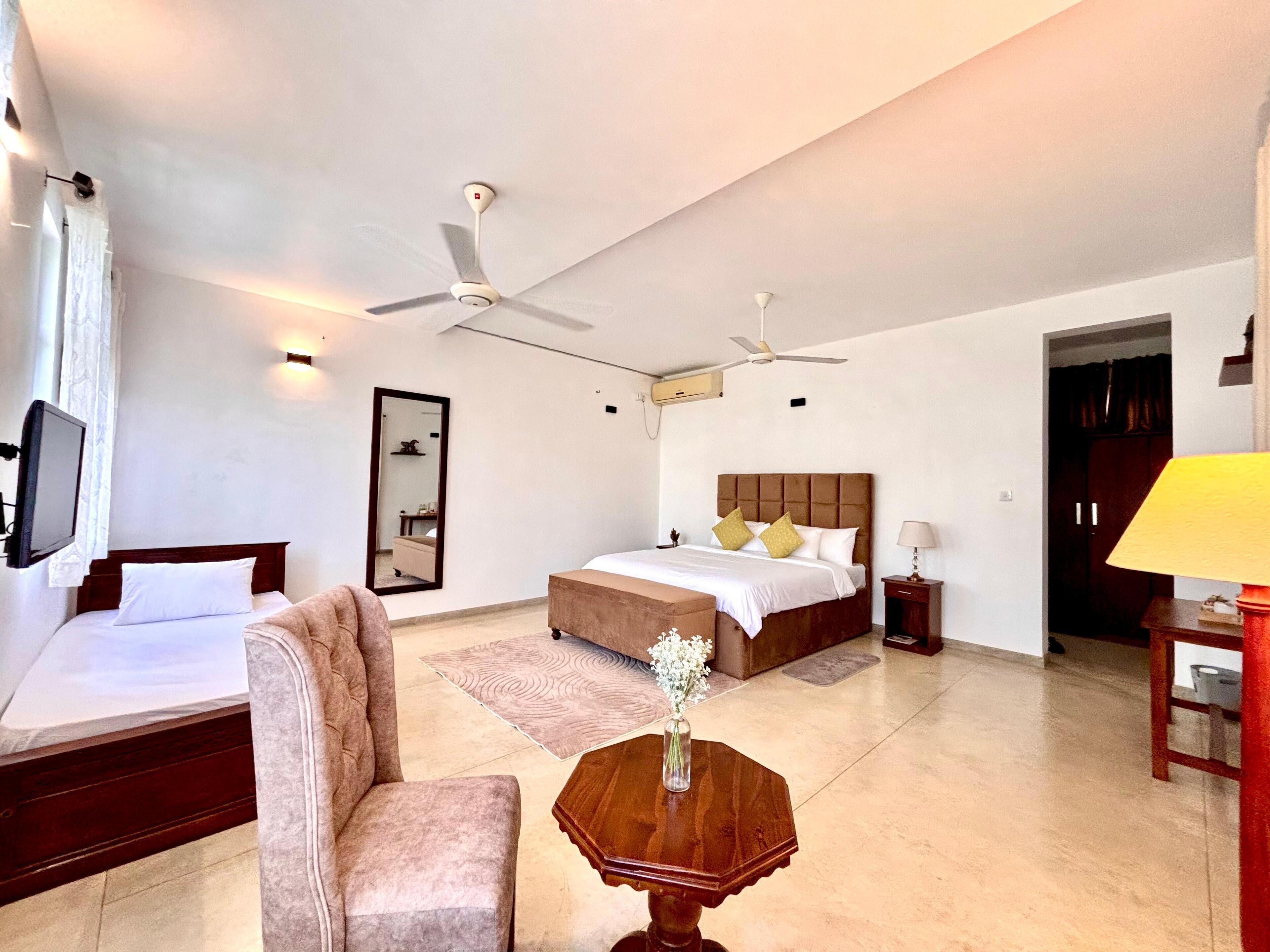 A hotel room with two beds, a TV, ceiling fans, a mirror, a bedside table, and a lamp.