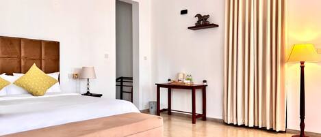 A hotel room with a bed, a bedside table, a lamp, a small table with items, a ceiling fan, and a window with curtains.
