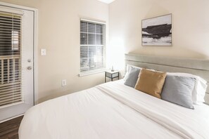 A bedroom with a large bed, a headboard, two windows with blinds, a bedside table, and a wall-mounted picture. - Cozy Retreat w/ Balcony & Modern Comforts (Birmingham)