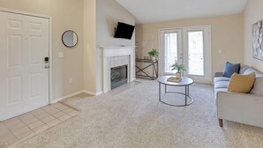 A living room with a fireplace, a flat-screen TV mounted above it, a round mirror on the wall, a sofa with cushions, and a small table with a plant. - Modern Comfort w/ Balcony, Kitchen & WiFi (Birmingham)