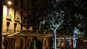An outdoor dining area with tables and umbrellas, surrounded by wooden planters and a building with balconies.