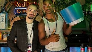 Two people standing in a bar area with a neon sign reading "Red Bull O P" and a bottle of liquor on the counter.