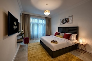Ein modernes Hotelzimmer mit einem großen Bett, einem Flachbildfernseher, einer Chandelüster und Blick ins Freie durch große Fenster.