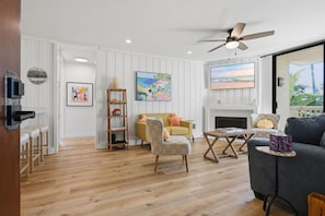 A spacious living room with a wooden floor, a ceiling fan, a fireplace, and a large window with a view of palm trees. - New Listing: Beautiful Interior with Two Balconies – North Coast Village G101 (Oceanside)