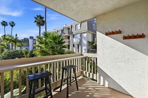 A balcony with a white railing, two black stools, and a view of a residential area with palm trees and buildings. - New Listing: Beautiful Interior with Two Balconies – North Coast Village G101 (Oceanside)