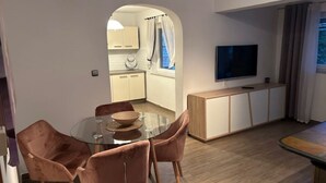 A modern living room with a glass dining table, four cushioned chairs, a flat-screen TV mounted on the wall, and a kitchen area visible through an archway. - Villa nikki, ideal base in croatia, 5o meters from the lake (Bilice)