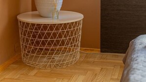 Una habitación con una mesita auxiliar redonda, blanca y de estructura metálica, que sostiene una lámpara blanca con pantalla blanca y un pequeño recipiente de vidrio con ramitas de junco.