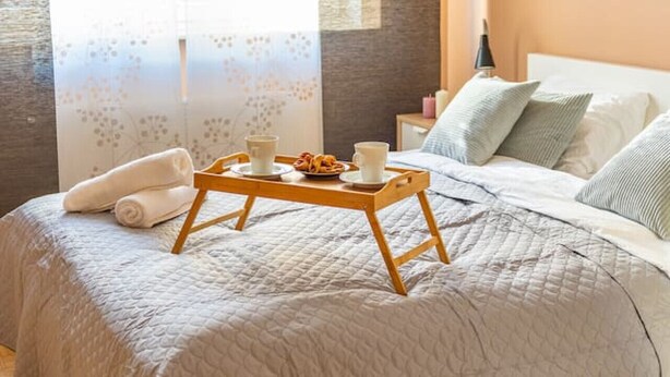 Una cama bien hecha con cubrecama acolchado, una bandeja de madera con artículos para el desayuno y una ventana con cortinas traslúcidas.