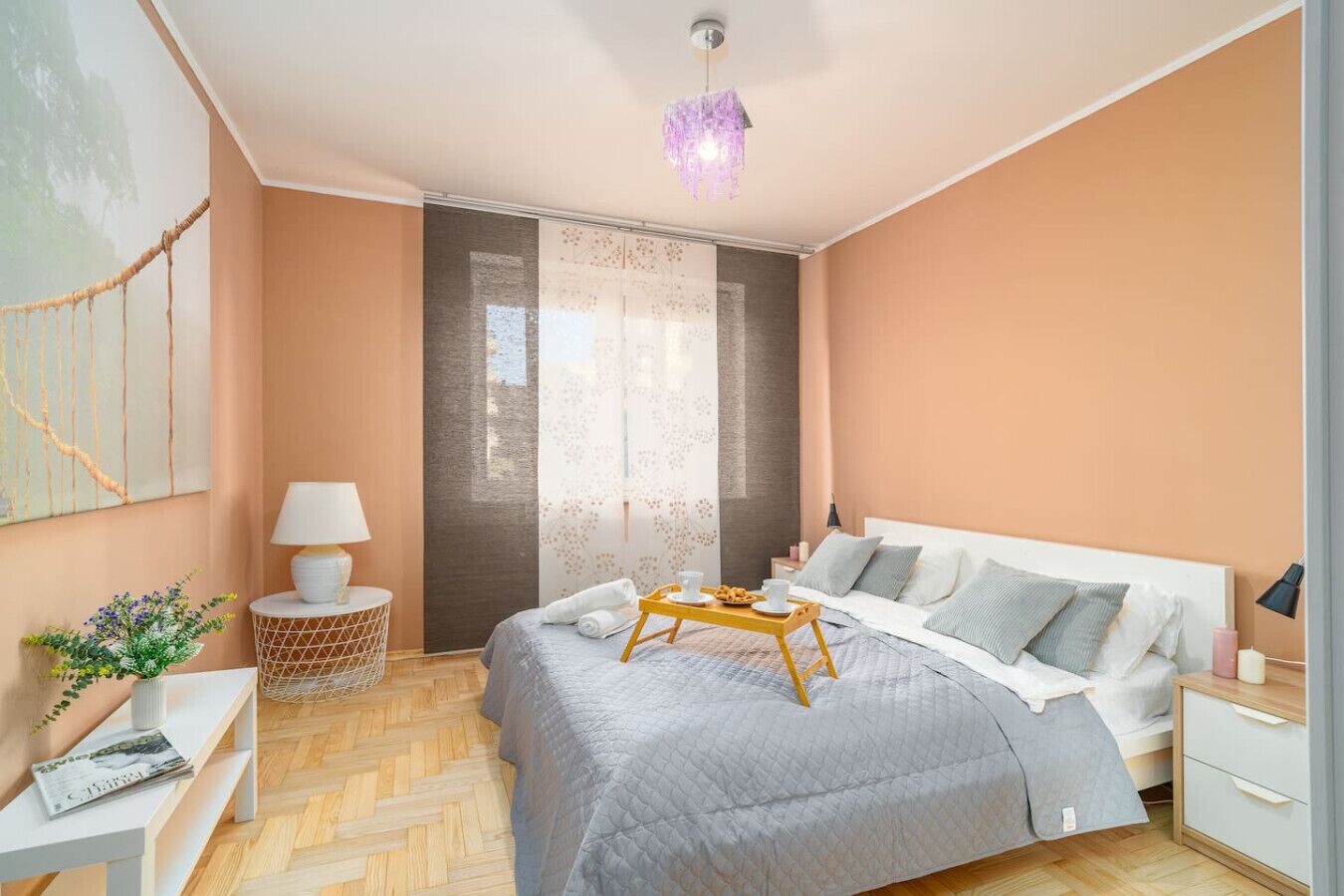 A modern bedroom with a large bed, a bedside table, a nightstand, a hanging light fixture, and a window with sheer curtains.