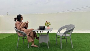 Une personne est assise à une table entourée de chaises, devant une petite table, à l’extérieur, sur une pelouse artificielle.