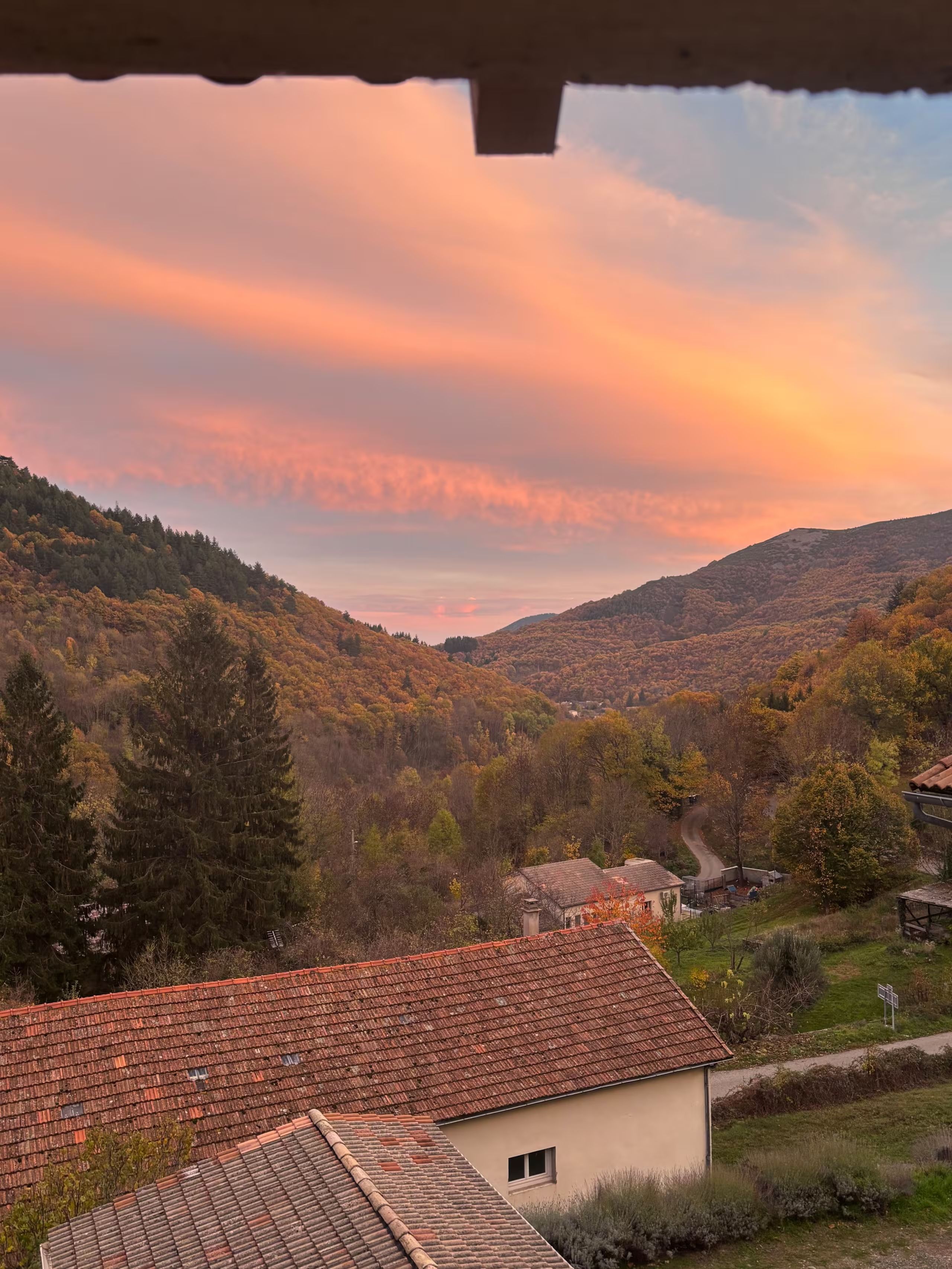 Un paysage rural avec une maison dotée d’un toit recouvert de tuiles rouges et d’une petite fenêtre, entourée d’arbres et de collines.