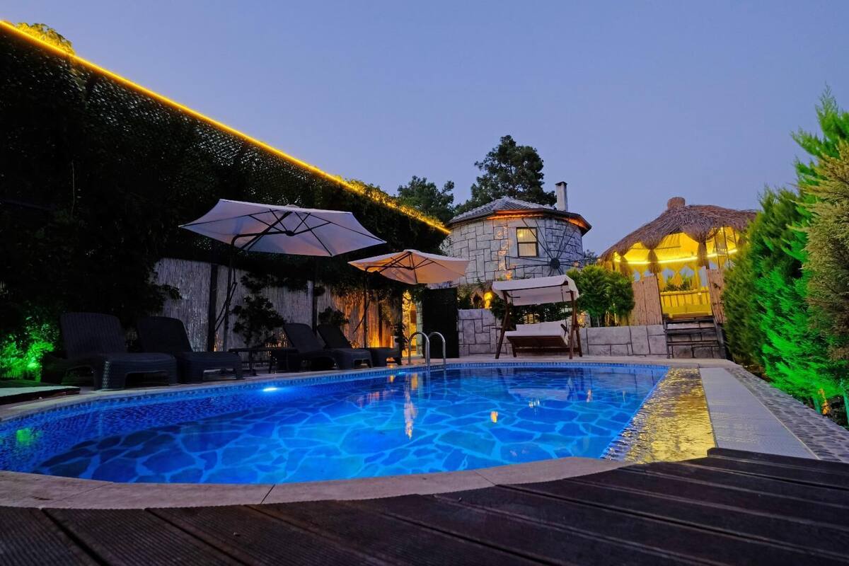 Una piscina con sillas de descanso y sombrillas, rodeada de muros de piedra y un edificio con techo de paja.