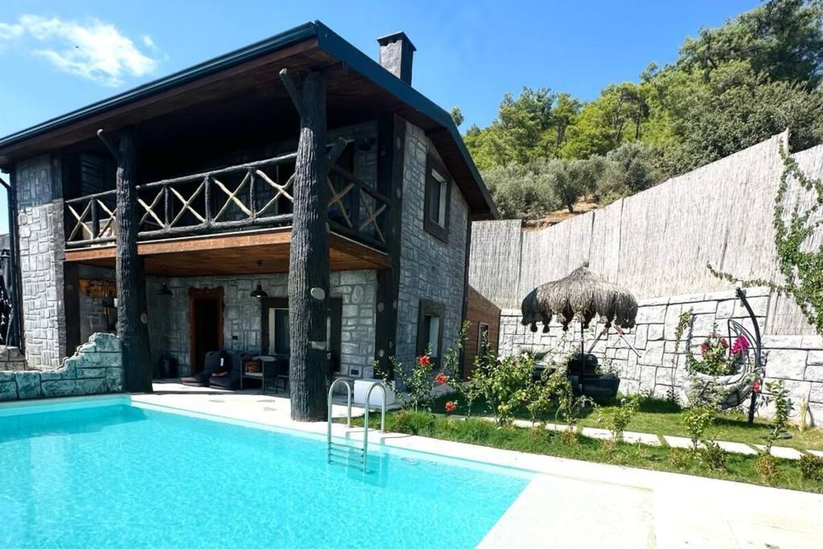 Un edificio de piedra con piscina, terraza de madera y un muro de piedra con techo de paja.
