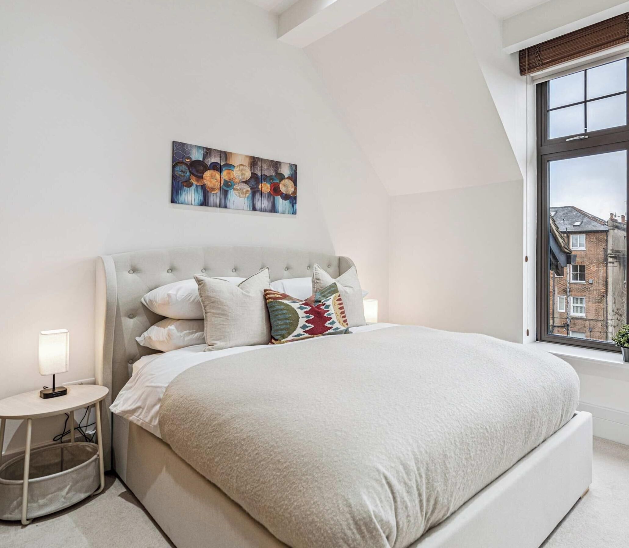 Une chambre avec un grand lit, une table de chevet avec une lampe, une fenêtre donnant sur des immeubles et une œuvre d’art accrochée au mur.