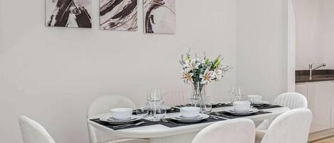 Et spiseområde med et rundt bord dækket til seks personer, hvide stole og en vase med blomster. Der er abstrakte malerier på væggen over bordet.